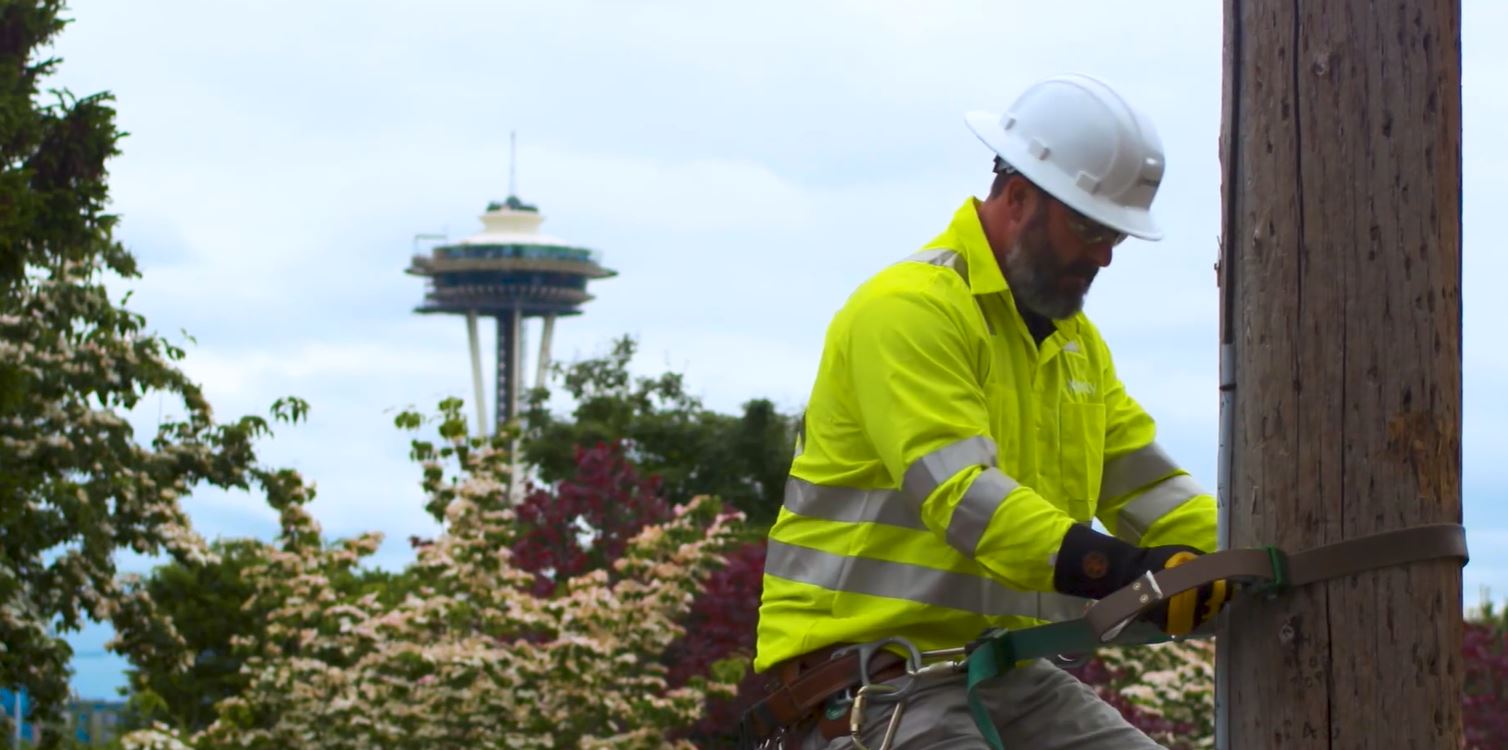 A Comcast technician works on a power pole with the Space Needle in the background.