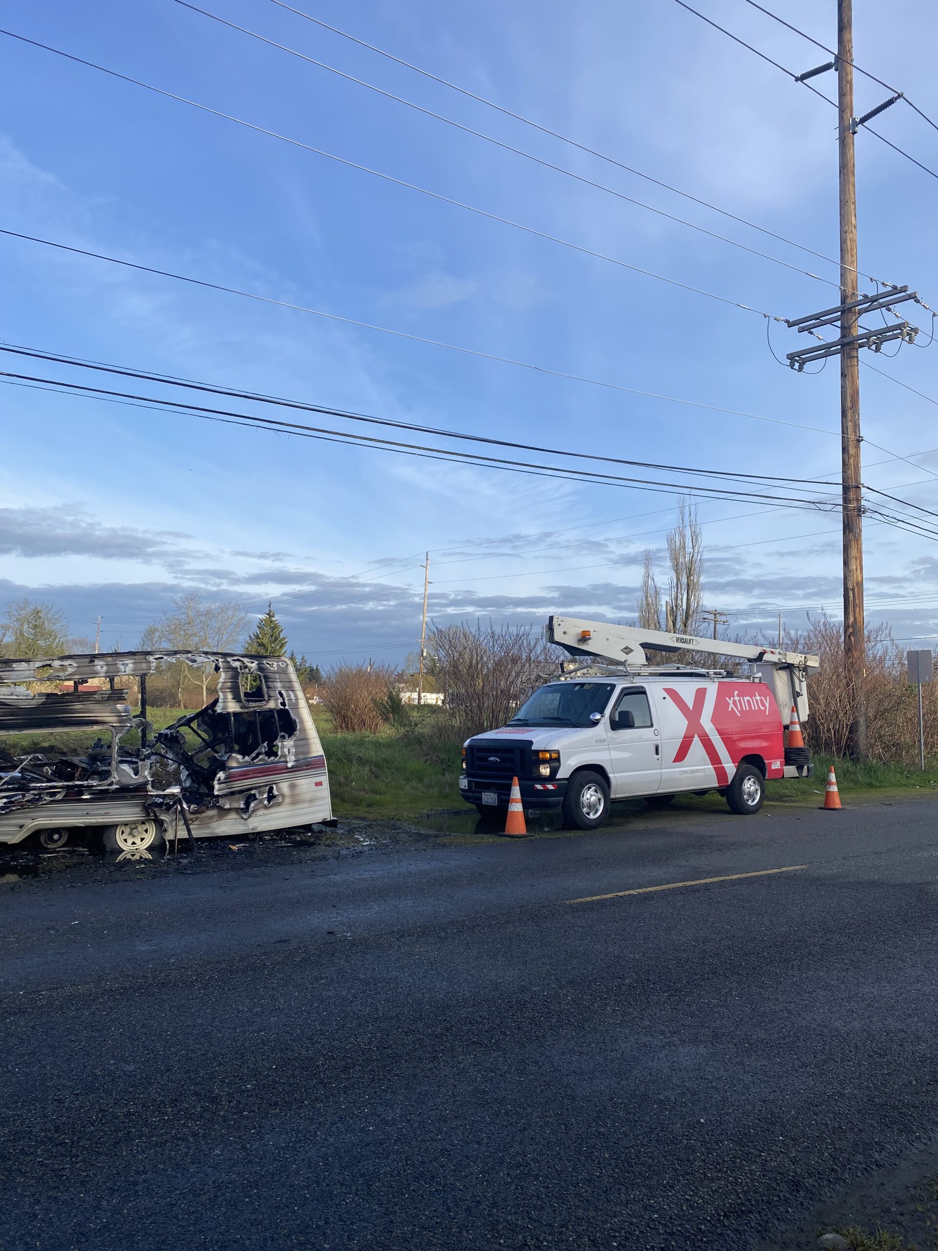 A burnt camper trailer next to an Xfinity van on the side of a road.