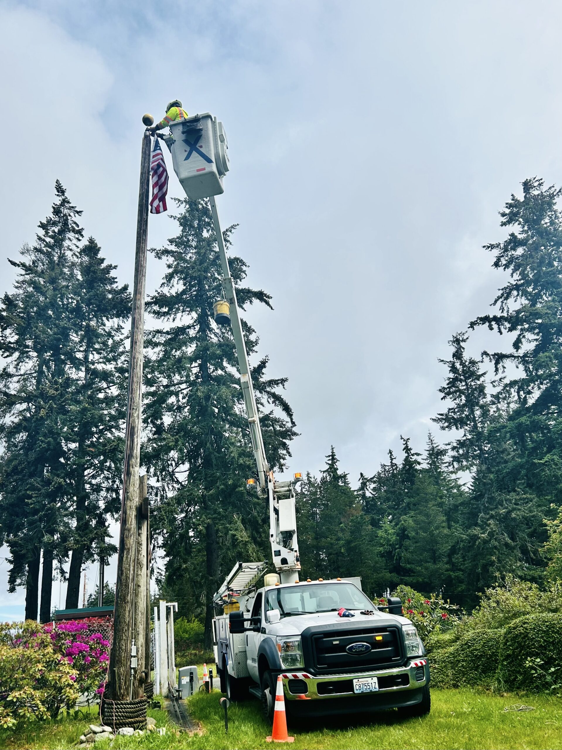 An Xfinity truck is parked by a flag pole and a Comcast technician repairs an American flag at the top of the pole.