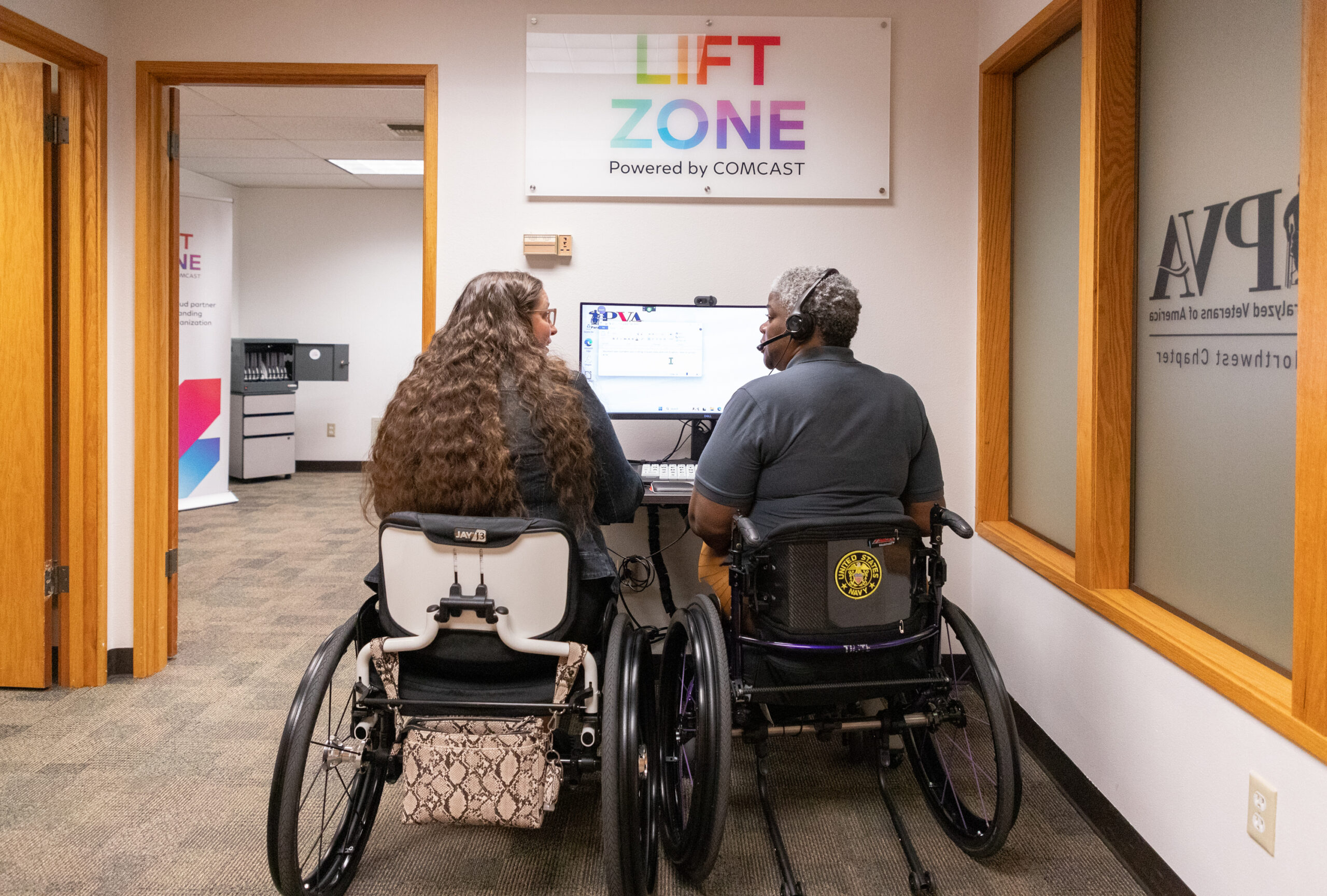 Two women in wheelchairs work at a computer under a Lift Zone sign.